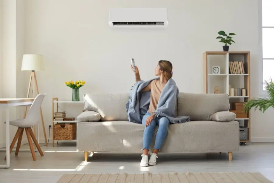 Woman seated on a beige sofa in a bright living room, holding a remote toward a wall-mounted white air conditioner above her.