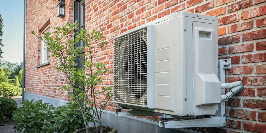 Outdoor air conditioner condenser unit mounted on a brick wall with a metal support bracket, nearby potted plant and shrubs in a sunny yard.