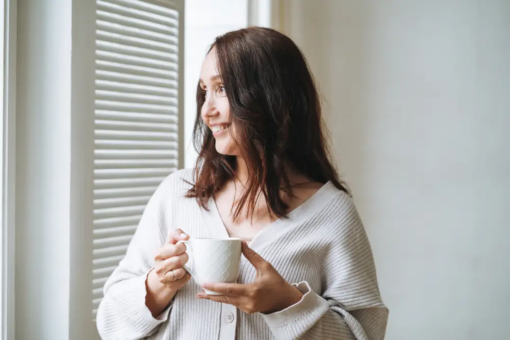 Furnace Tune-Up Benefits in Auburn, AL - Young woman in cozy knitted cardigan with cup of tea in hands in bright interior at home