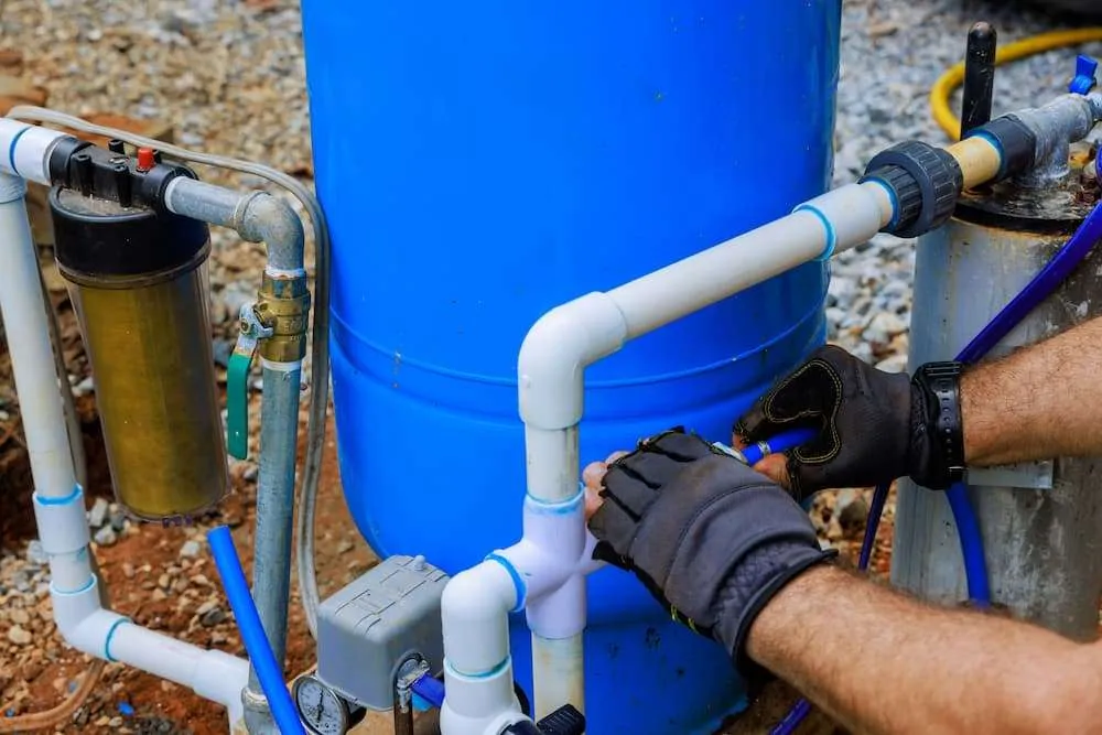 Close-up of hands in work gloves repairing PVC piping and a sediment filter attached to a large blue outdoor water pressure tank