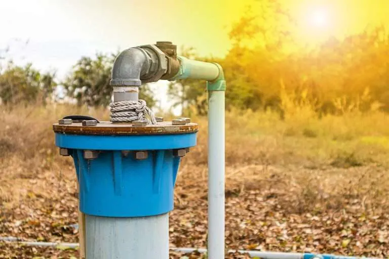 outdoor blue residential well water head assembly with a metal cap and PVC pipe connection in a field at sunset