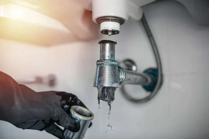 close-up of a plumber's gloved hand working on a leaking silver P-trap assembly under a white porcelain kitchen sink