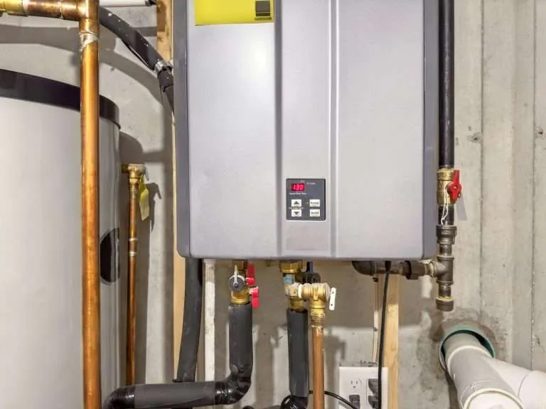 silver wall-mounted tankless water heater with a digital display showing 130 degrees, connected to insulated copper pipes and a gas line