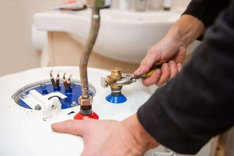 Close-up of a plumber's hands using a wrench to tighten brass fittings on top of a white residential water heater in a bathroom