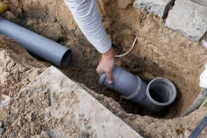 worker's hand connecting a grey PVC pipe fitting into an underground sewer line trench during a professional drainage repair
