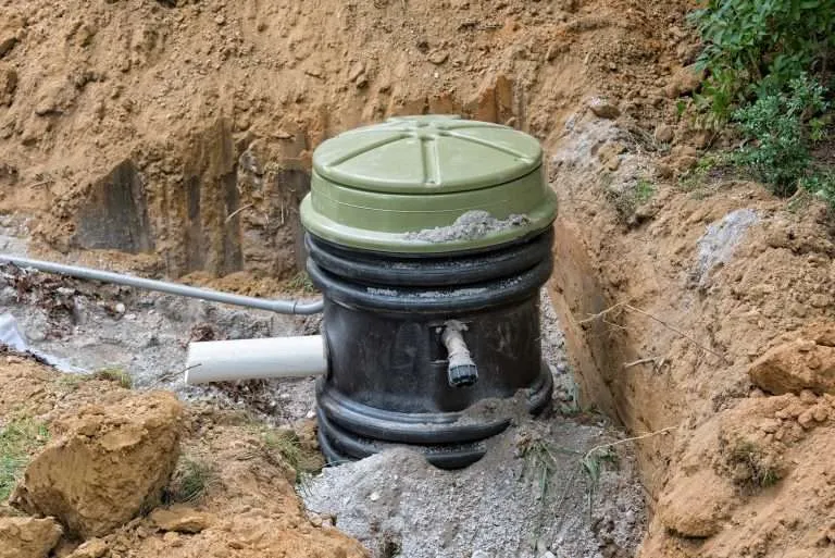 A newly installed black plastic underground septic tank riser with a green lid, positioned in an excavated dirt trench with connecting white PVC pipes