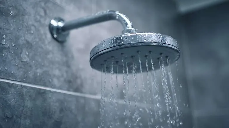 side-view of a large circular chrome rain shower head with water streaming from multiple nozzles against a grey tiled wall
