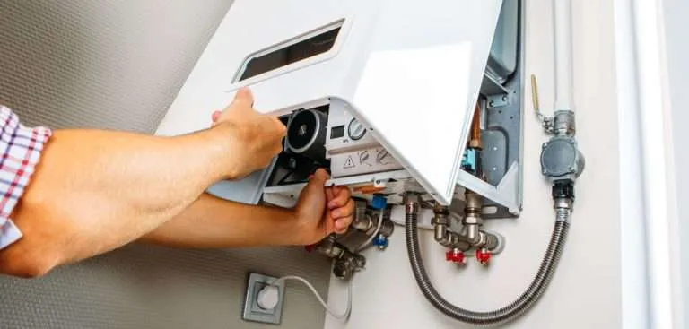 close-up view of a technician's hands opening the front panel of a white wall-mounted tankless water heater for maintenance or repair