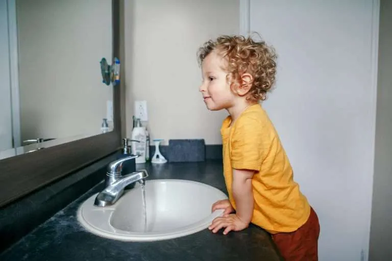 young toddler with curly hair standing at a bathroom vanity, looking curiously at a stream of water flowing from a chrome faucet into a white porcelain sink