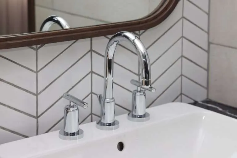 modern polished chrome three-piece bathroom faucet installed on a white sink against a white herringbone patterned tile backsplash