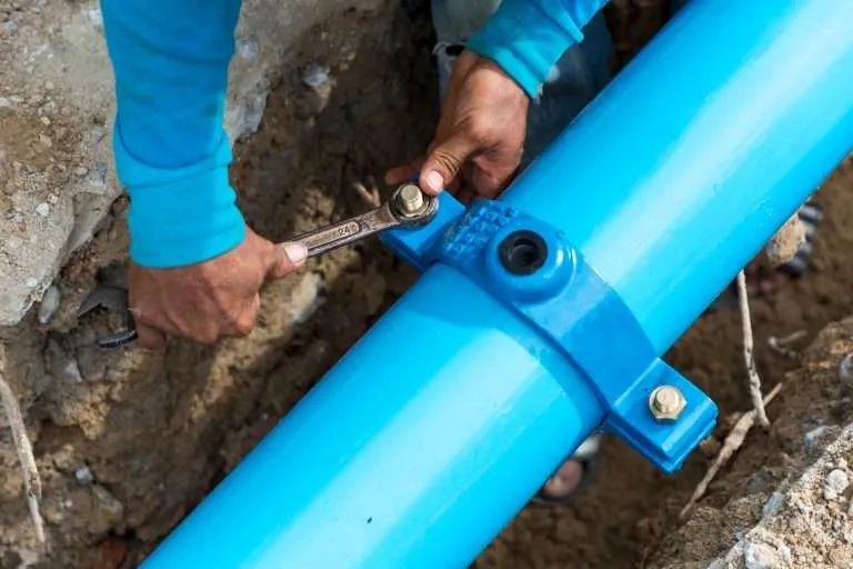 worker using a large wrench to tighten a heavy-duty blue coupling on a thick blue underground water main pipe in a trench