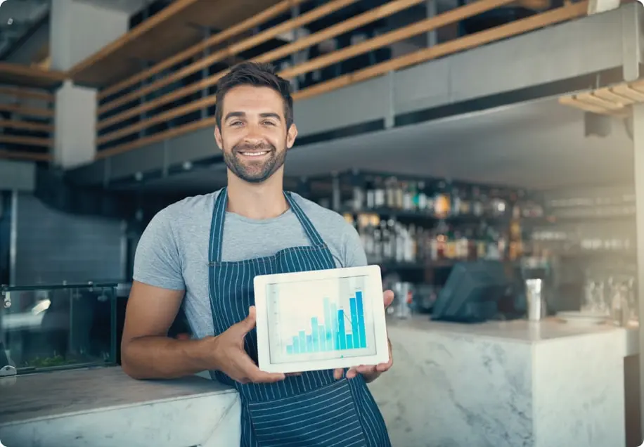 A smiling man wearing an apron stands holding a tablet showing a bar graph.