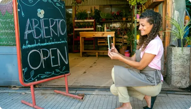 A smiling female business owner wearing an apron takes a photo of her store's open sign