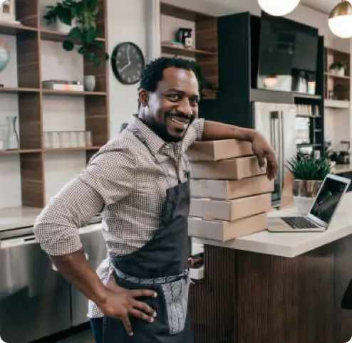 A smiling male business owner wearing an apron rests his arm on a stack of boxes