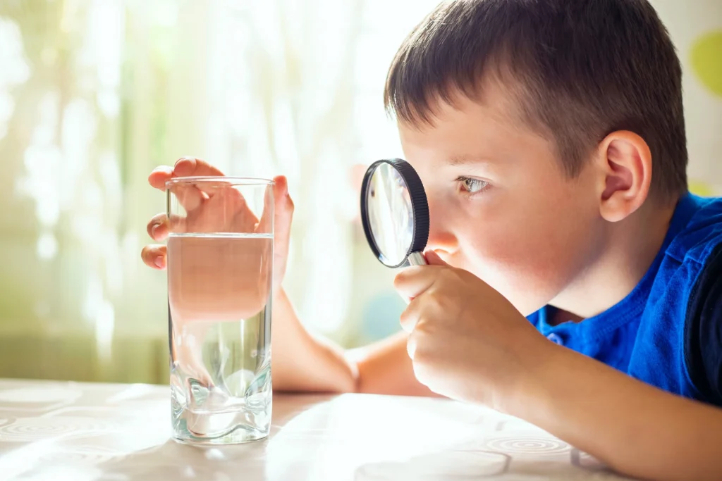 Boy looking through a magnifying glass at a glass of water on a table.