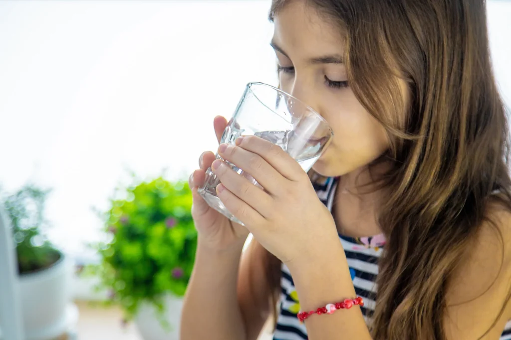 Young girl drinking a glass of water indoors, with green plants in the background.