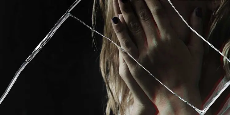 Close-up of a distressed woman holding her face in hands, viewed through cracked glass symbolizing trauma and legal reporting.