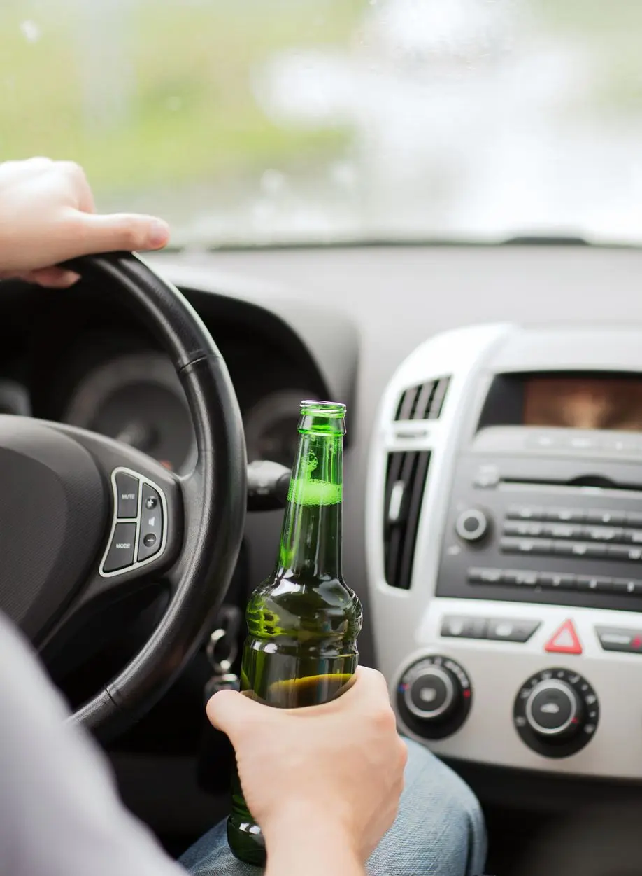 A driver holds a green glass beer bottle in one hand while gripping a car’s steering wheel with the other.