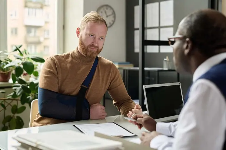 Injured client with an arm sling discussing a personal injury claim with an attorney at an office desk.
