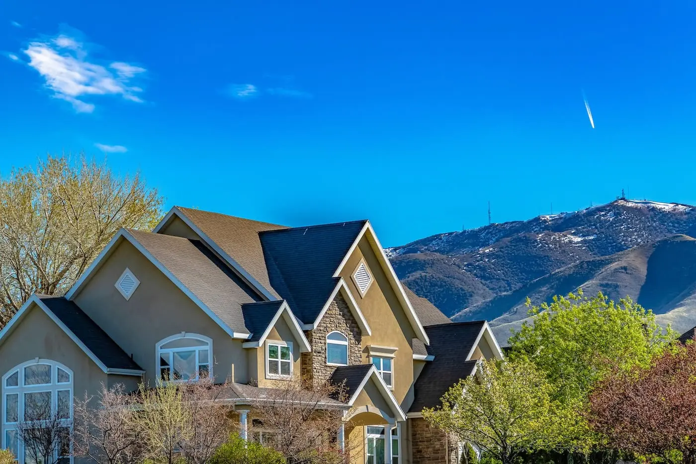 House exterior against mountain and blue sky viewed on a sunny day. The lush trees and outdoor scenery are reflected on the windows with white frames.