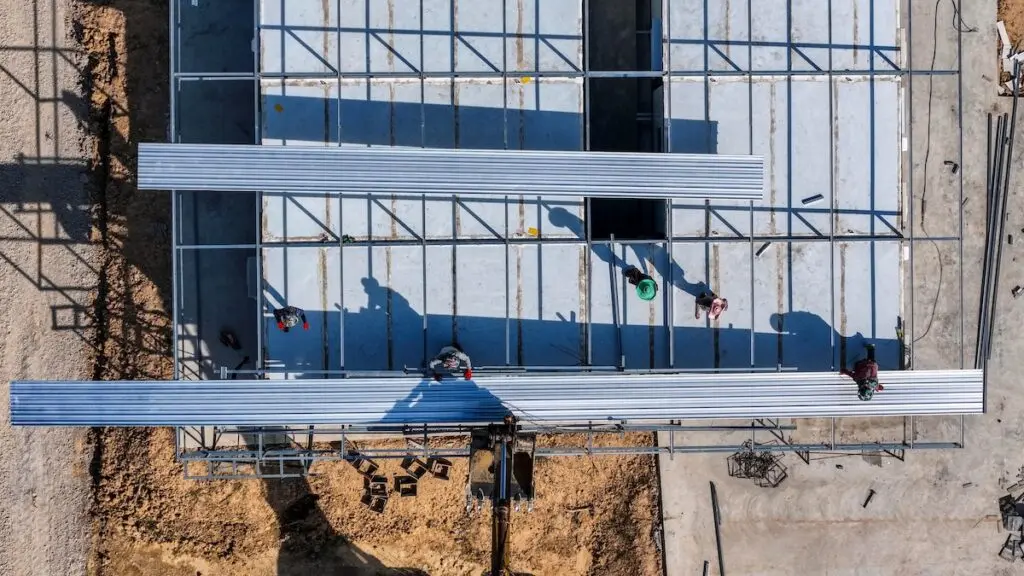 Aerial view of workers placing long metal sheets onto a building framework with the aid of heavy machinery, highlighting precision, teamwork, and modern techniques in construction projects.