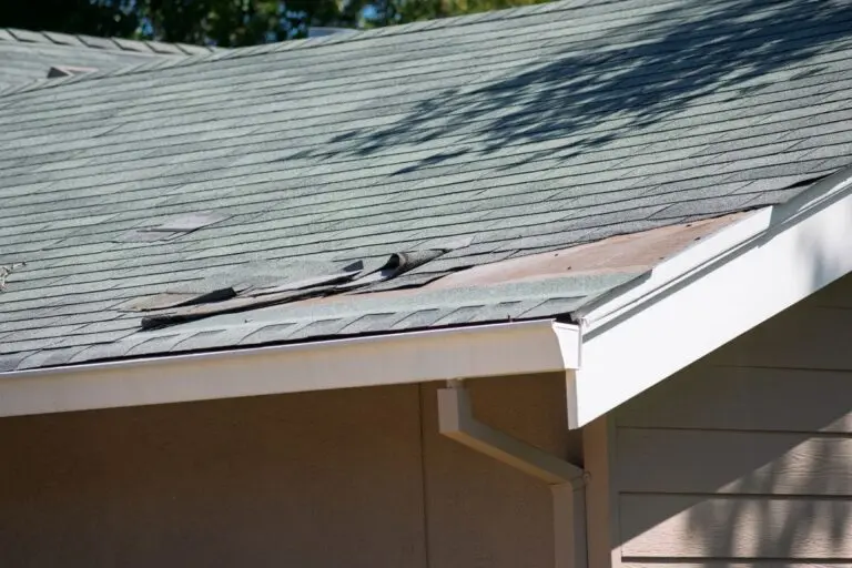 Damaged asphalt shingles are visible on a residential roof, with several pieces curled and missing, revealing the wooden underlayment beneath
