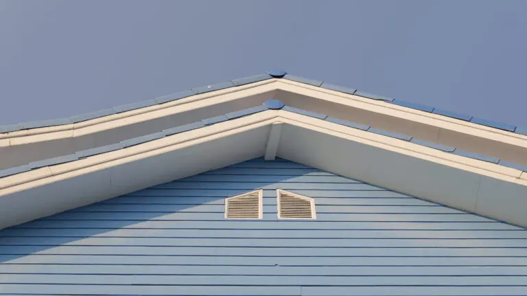 A low-angle architectural shot showcases the clean lines of a blue wooden house gable featuring white trim and symmetrical ventilation slats under a clear sky