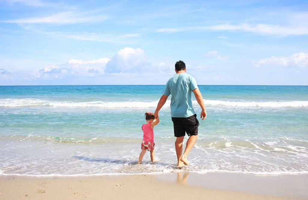 A father and daughter hold hands while walking on a sunny beach, enjoying family time together.