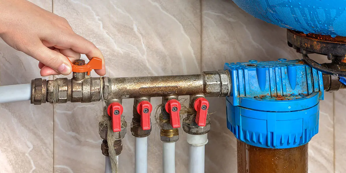 Hand turning a red valve handle on a main plumbing pipe manifold to shut off water during a household emergency.