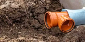 An orange PVC sewer pipe connector and T-fitting sit inside an excavated trench during sewer line replacement.