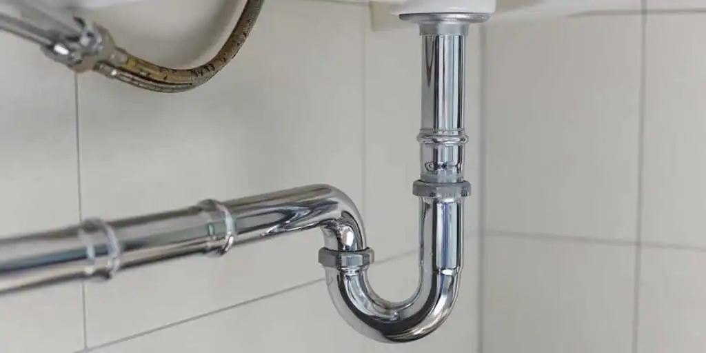 A polished chrome P-trap connects the drainage pipes beneath a bathroom sink against a white tiled wall.