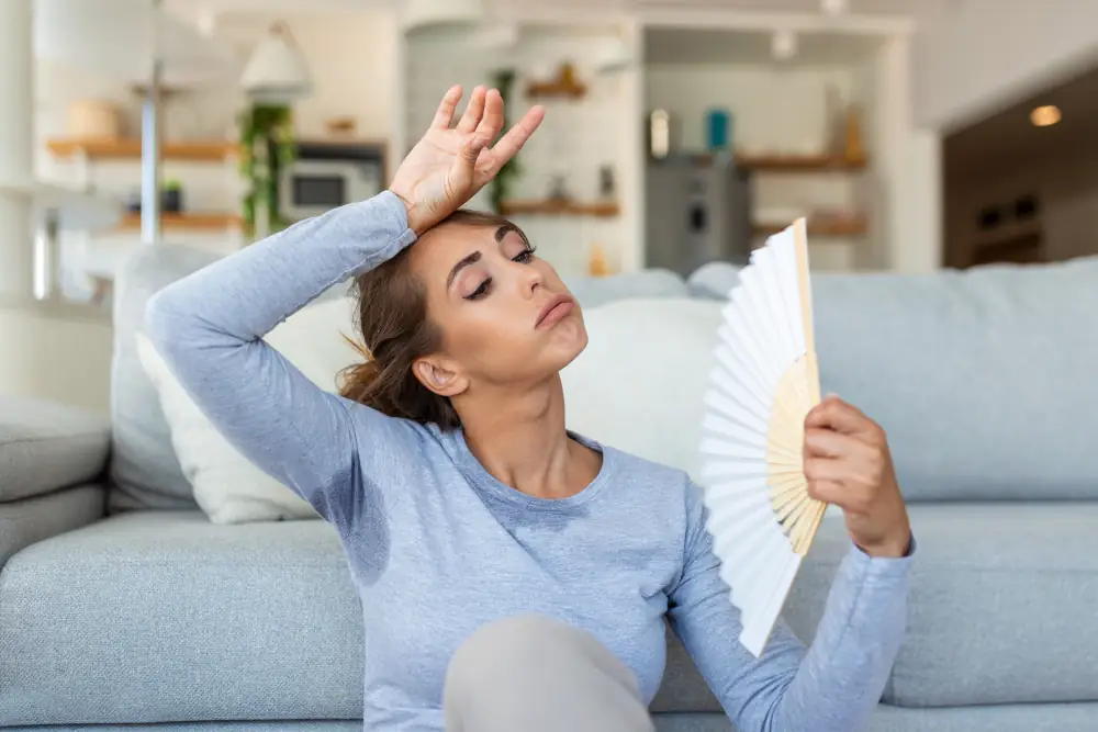 The Telltale Signs: Recognizing When Your AC System Requires Repair or Replacement - Woman sitting on the floor in front of her sofa. She is sweating and holding a paper fan with her left hand