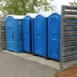Row of blue portable toilets, including an ADA-compliant unit, installed in a public park area with privacy fencing for events or high-traffic outdoor spaces