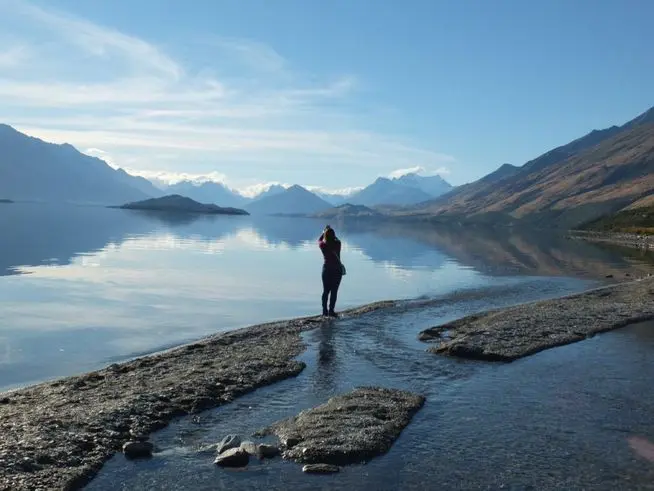 Person standing by calm lake with mountains