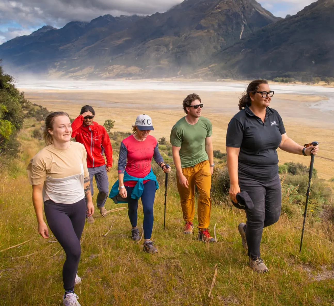 Guide leading a small group of walkers up a grassy slope above a wide braided river valley
