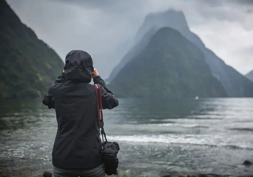 Visitor in a rain jacket photographing Mitre Peak in cloud at Milford Sound
