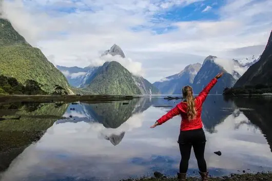 Visitor with arms outstretched in front of Mitre Peak reflected at Milford Sound