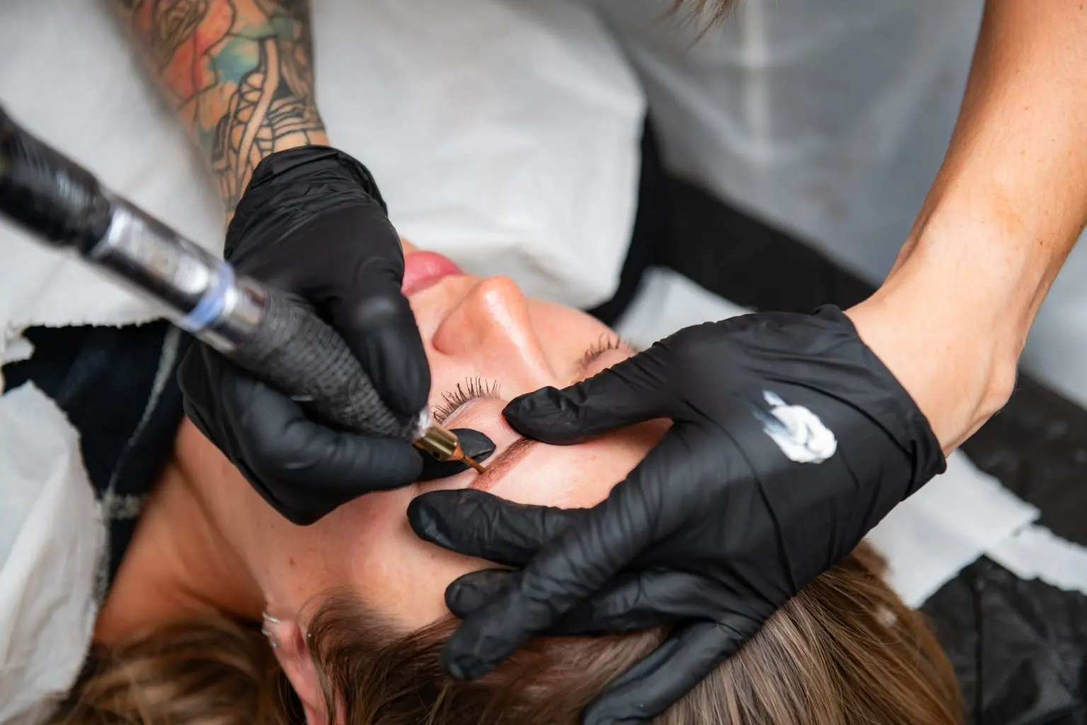 Gloved hands holding a permanent makeup machine against a model's brow during the live competition