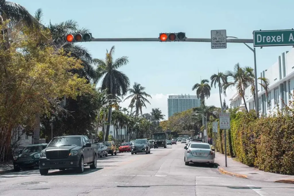 Vehicles driving down a palm-tree-lined coastal street toward a high-rise under red traffic signals.