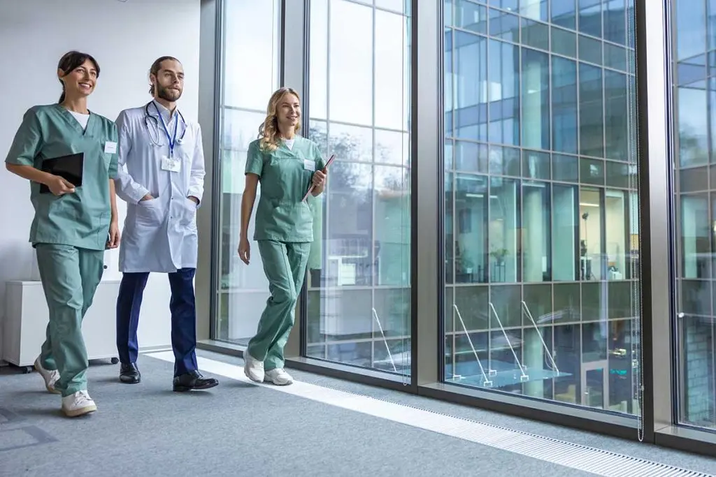 Three smiling healthcare professionals in scrubs and lab coat walk down a bright corridor past large floor-to-ceiling windows.