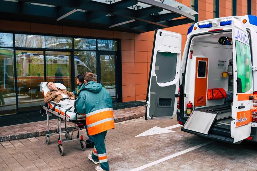 Paramedics push a patient lying on a stretcher toward an open ambulance parked right outside a modern hospital entrance.