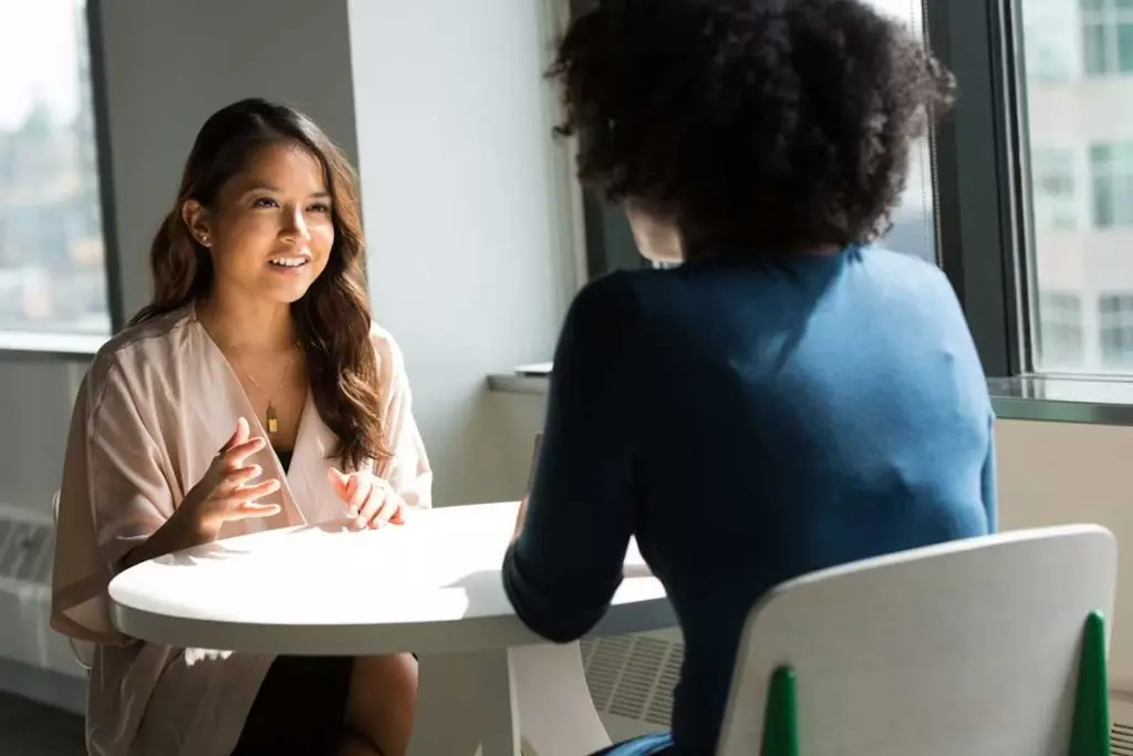 Two women having a professional conversation at a table in a sunlit office space.