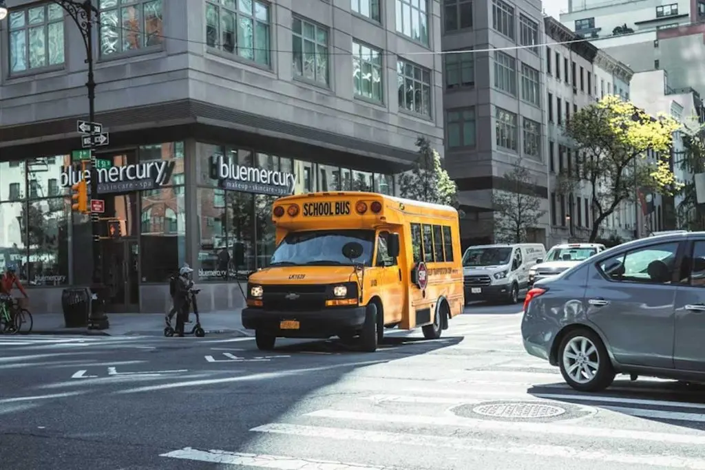School bus turning through an intersection alongside a sedan, delivery van, cyclist, and scooter rider.