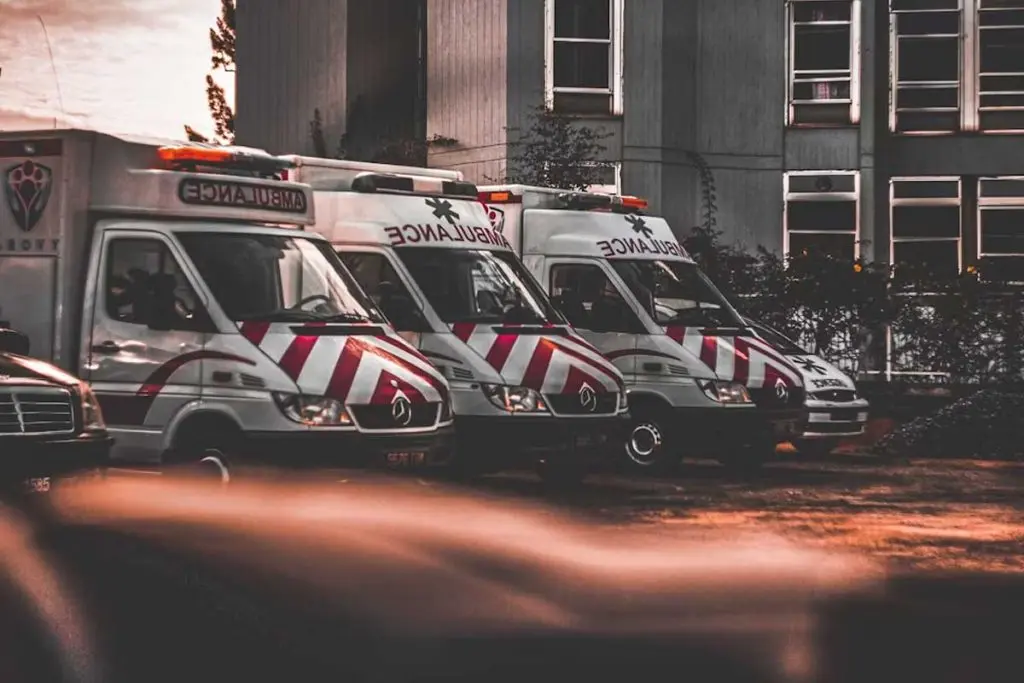 Row of parked ambulances with chevron striping outside a multi-story facility under low-light skies.