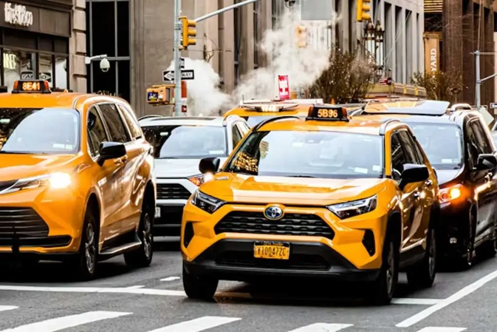 Yellow taxicabs navigating a crowded city street past steam rising from a utility vent near storefronts.