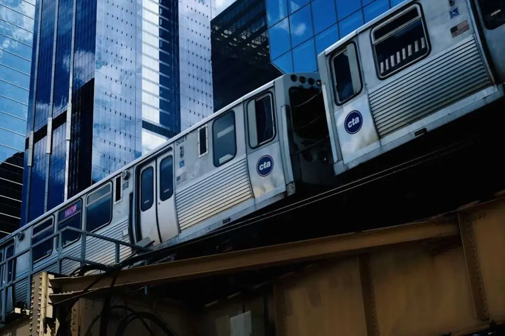 Elevated subway train moving along an outdoor rail structure past reflective glass towers.