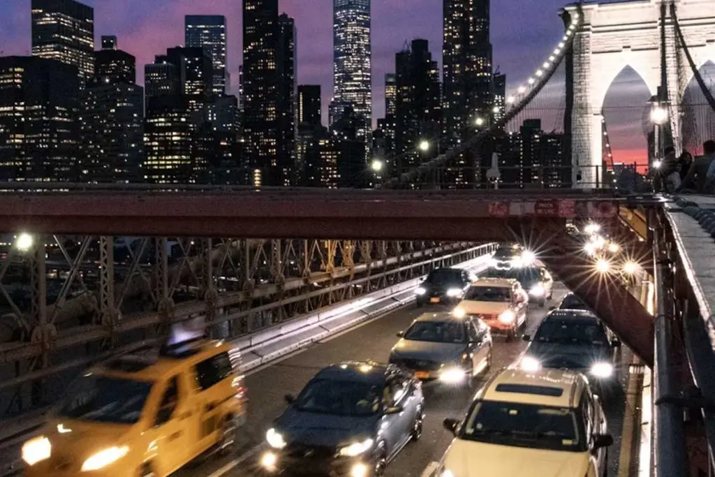 Cars streaming across a bridge roadway at dusk with a lit gothic stone tower and city skyline behind.