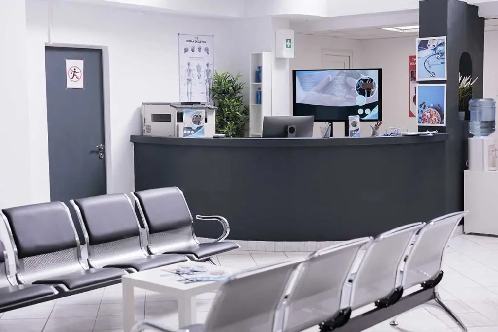 Modern medical center waiting room with rows of empty chairs, a clean reception desk, display screens, and educational posters.
