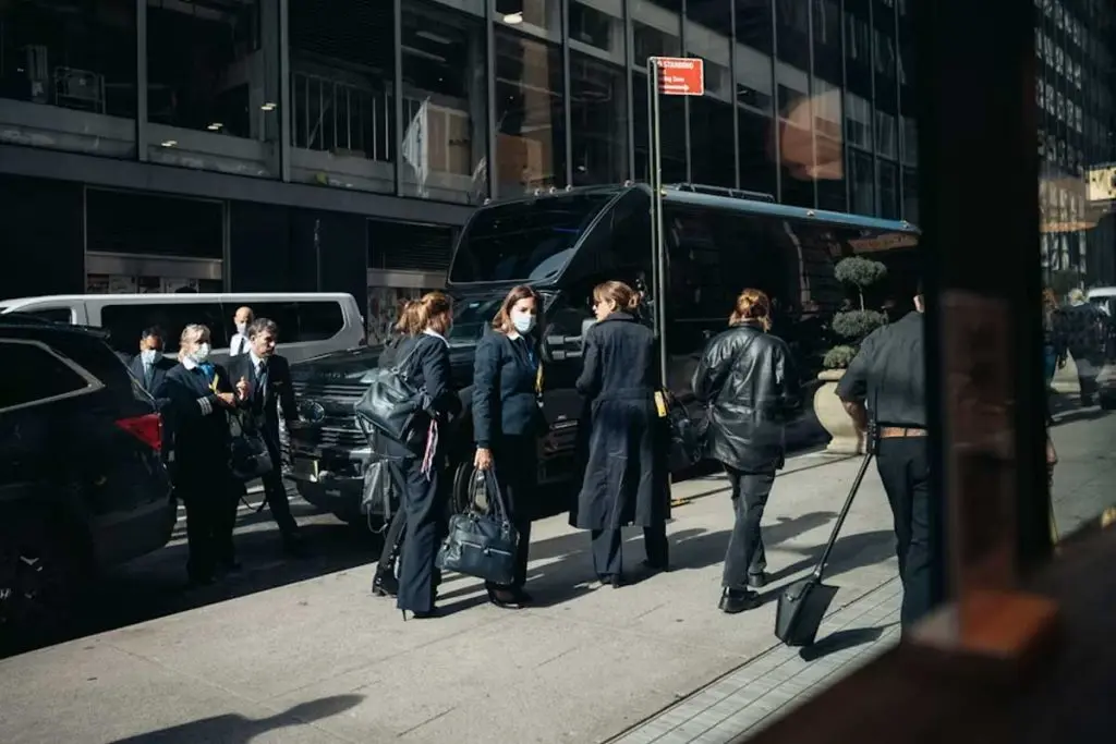 Flight crew members gathered on a sunlit sidewalk beside a parked shuttle van and executive SUVs.
