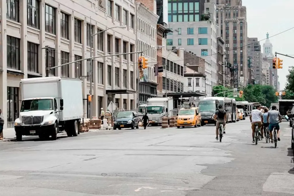 Cyclists riding along a wide avenue past parked delivery trucks, vans, and a taxi under midday light.